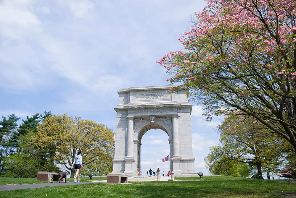 A large white arch in Valley Forge National Park in Philadelphia's Countryside stands tall in the middle of a park. To the left, a woman walks her dog on a trail. To the right, there is a large tree with pink blossoms. Through the arch in the distance, there are numerous people shown walking around.