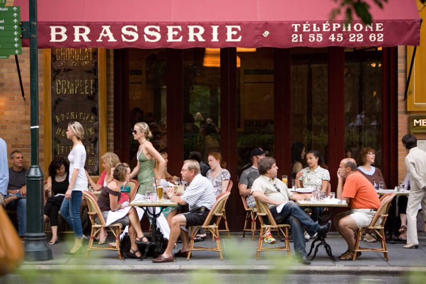 Two women are shown walking past tables of people sitting enjoying a meal on the sidewalk outside of Parc. The canopy overhead is maroon with white writing reading Brasserie.