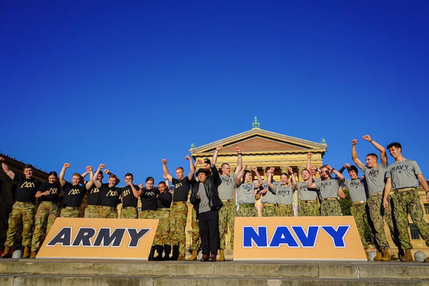 There are two groups posing for a picture in front of the Philadelphia Museum of Art. On the left, there is a group representing the Army behind a large sign that reads Army in all capital letters. On the right, there is a group of people representing the Navy behind a sign that reads Navy in all capital letters. In the middle, a Rocky impersonator stands with his fist in the air. Some of the individuals on either side are also holding their fists in the air.