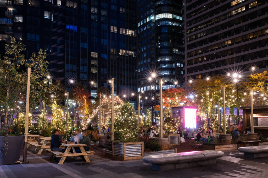 People sitting at picnic tables surrounded by holiday trees with lights and rustic décor surrounded by nearby skyscrapers