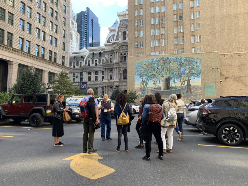 A group of people are shown standing in a parking lot, listening to a tour guide as he talks about the mural in front of them. There is a large mural depicting a tree with people shown climbing ladders around the tree. The mural appears to be on the side of a building to the right of Philadelphia City Hall, a large building.