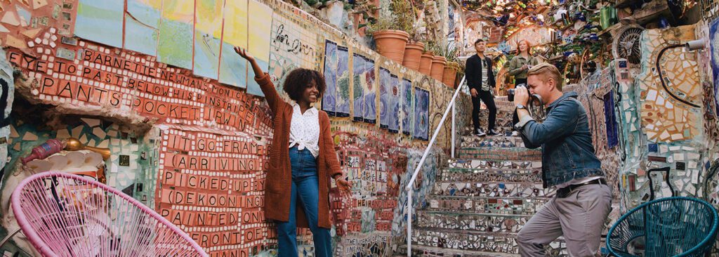 A person poses in front of a color mosaic wall while another person takes a photo.