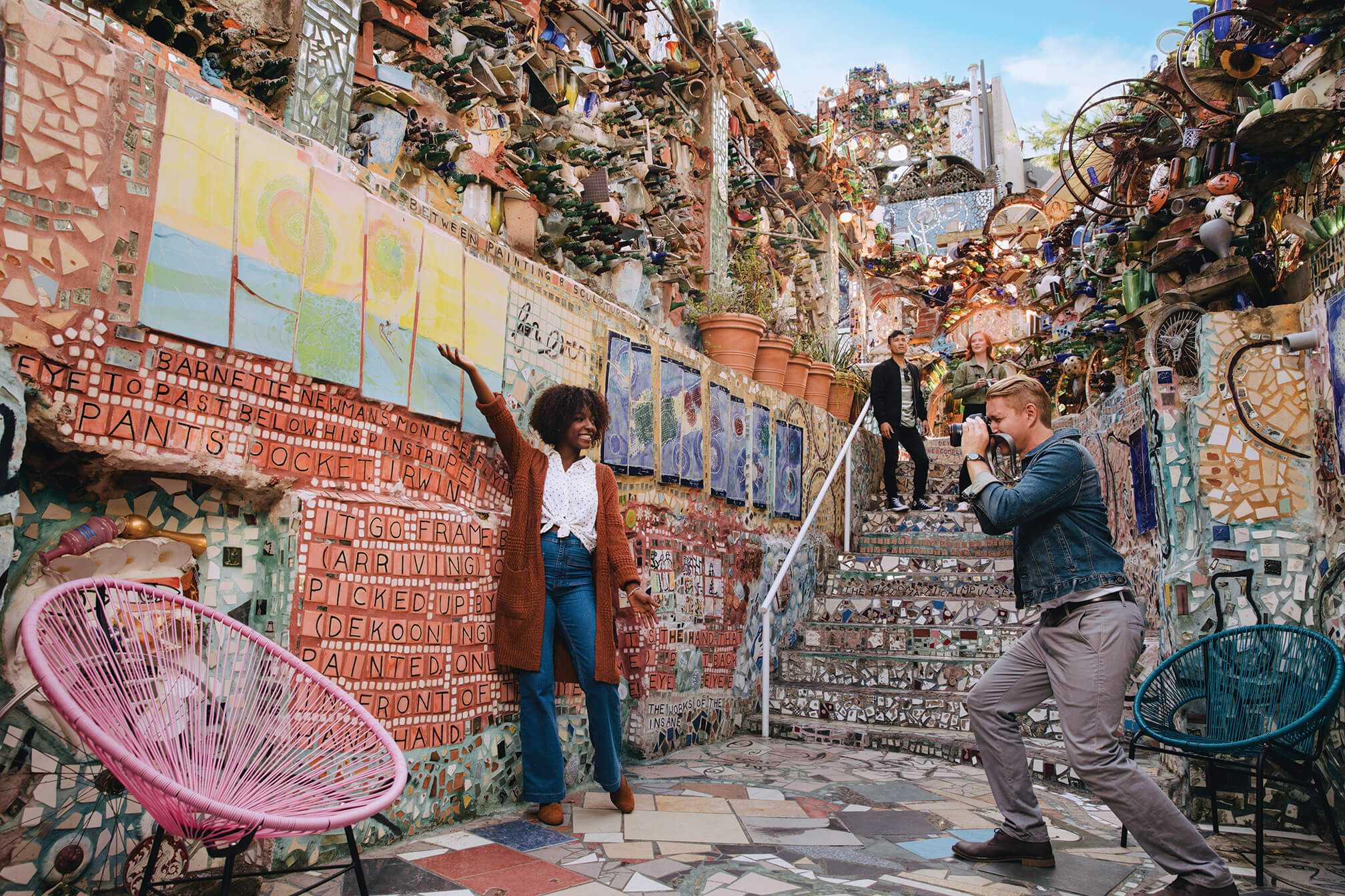 A person poses in front of a color mosaic wall while another person takes a photo.