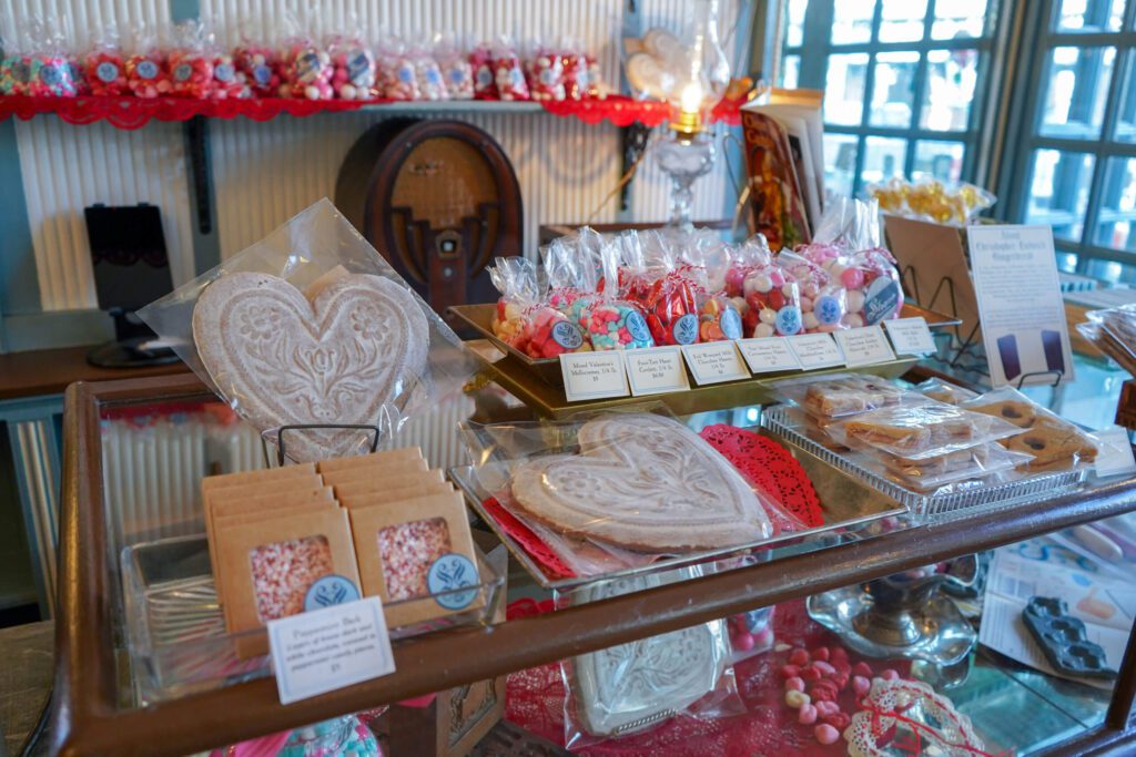 Heart-shaped candies and sweets are shown on top of a glass cabinet inside of a classic candy store. Red decorations fill the store in celebration of Valentine's Day.