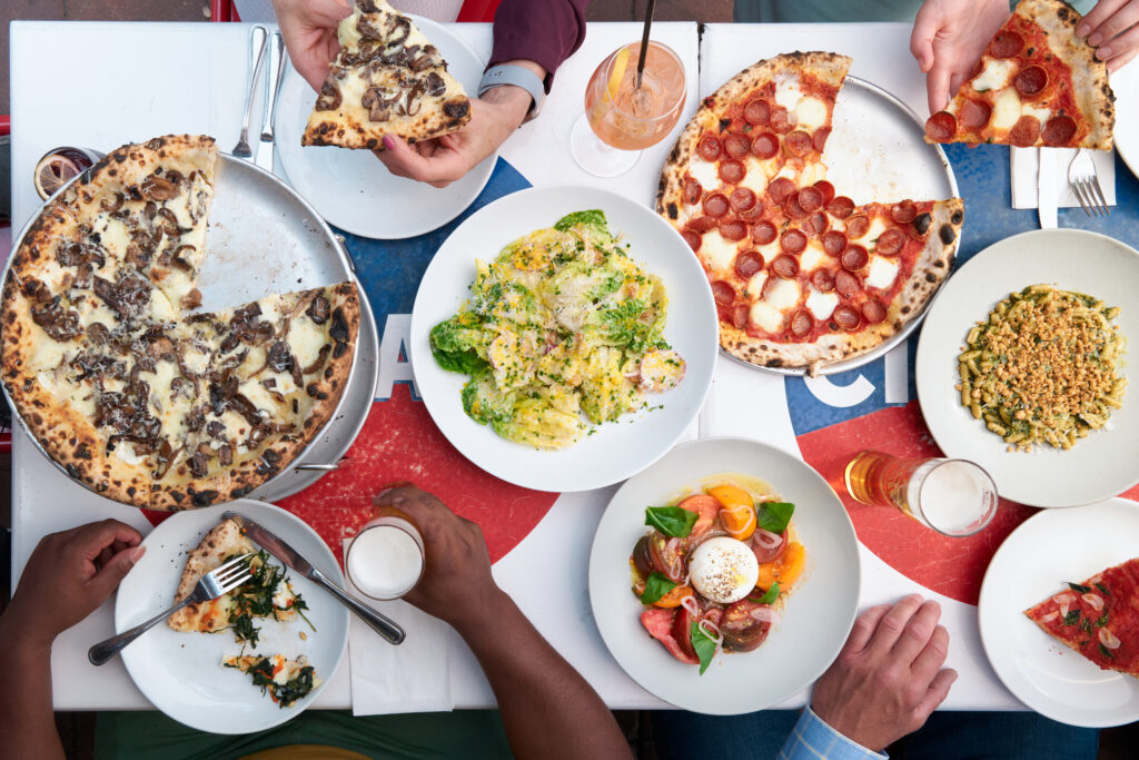 A table of food from Pizzeria Stella, including a pepperoni pizza, mushroom pizza, and a pasta plate.