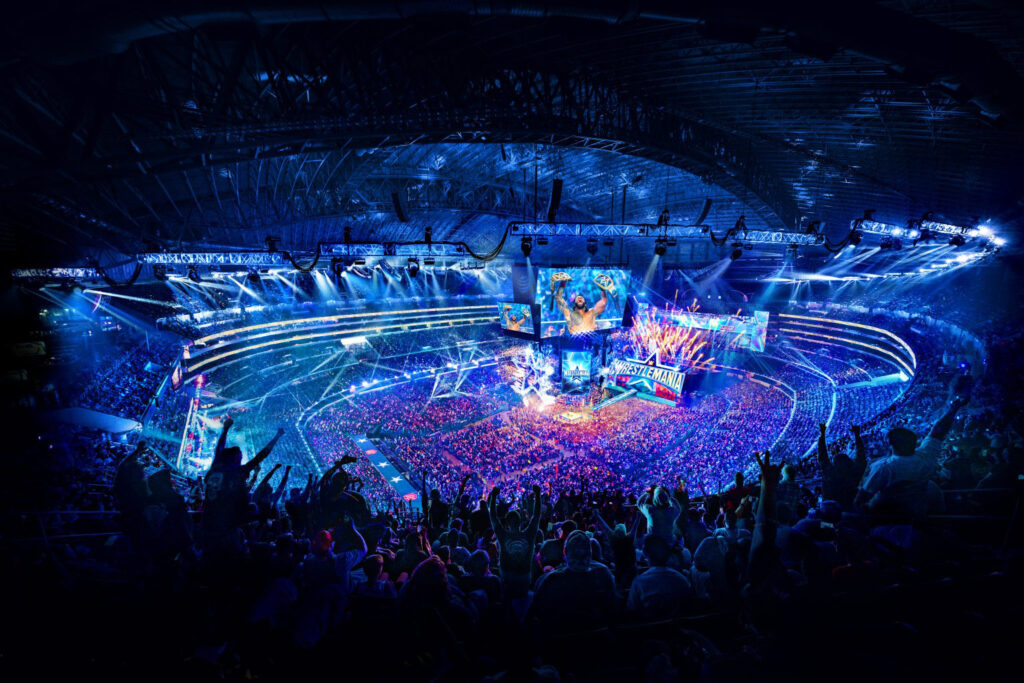 A stadium is shown lit up in blue lights. Massive screens are showing a Superstar holding up two belts. Fans cheer from their seats, some have their hands and arms in the air.
