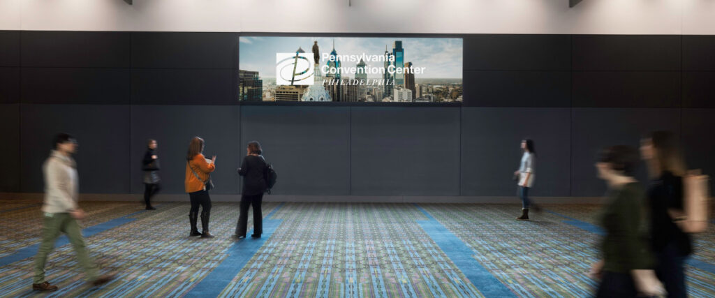 People moving quickly around a carpeted room with a television screen in the background.