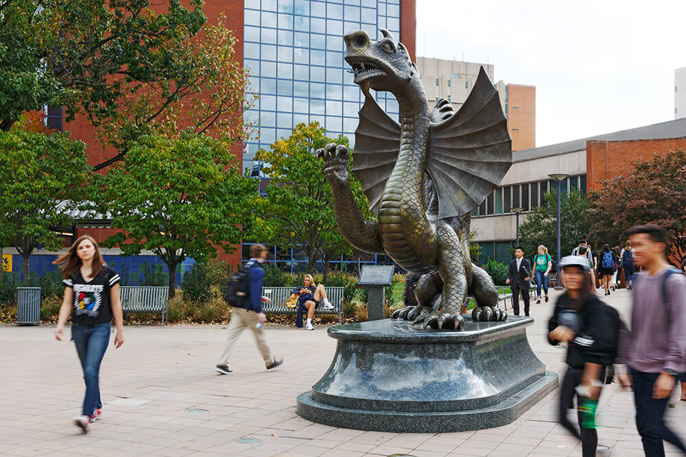 A large sculpture of a dragon is shown. There are students shown walking on either side of the statue. In the background, there is a large building made of bricks and glass. The windows reflect the sky.