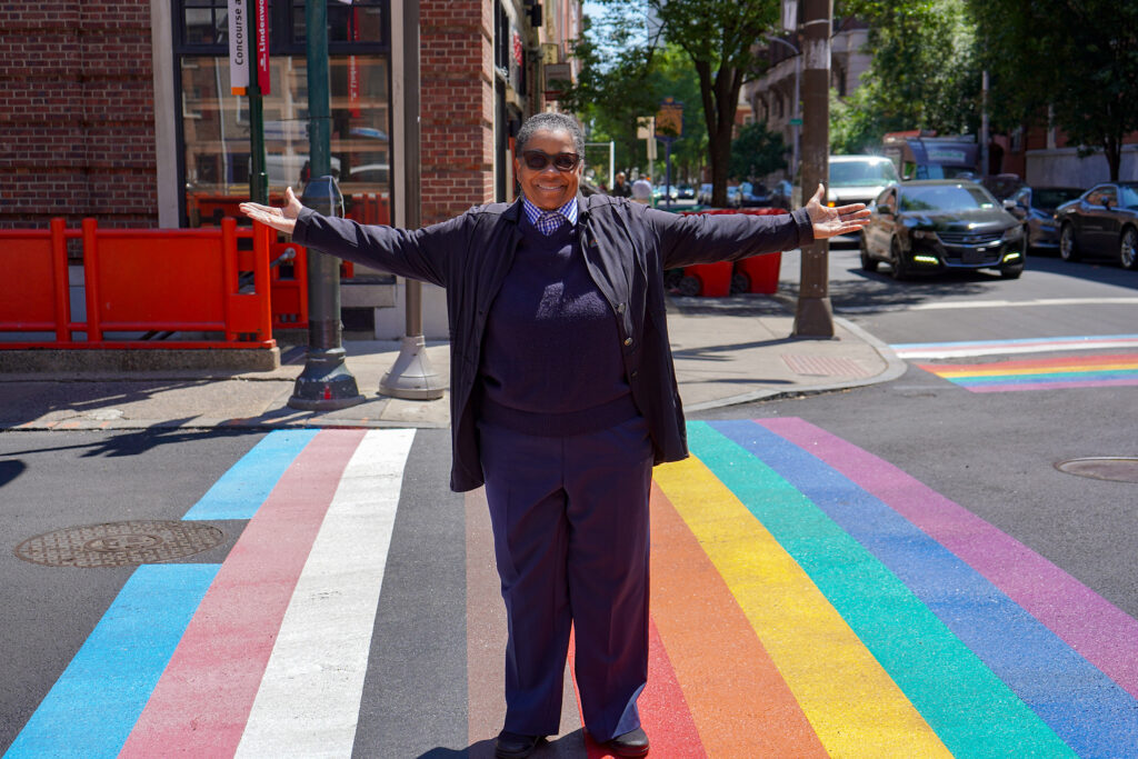 A person stands in the middle of a rainbow crosswalk with their arms out.