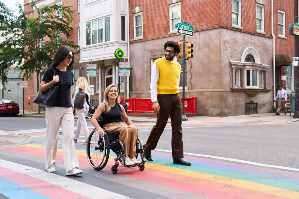 A man, a woman, and a woman in a wheelchair cross a rainbow painted crosswalk in Philadelphia's Gayborhood.
