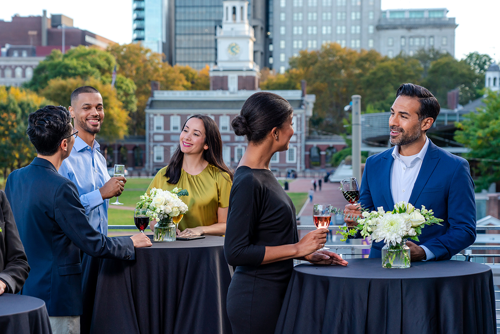 A group of people enjoying cocktails on a rooftop with Independence Hall in the Background.