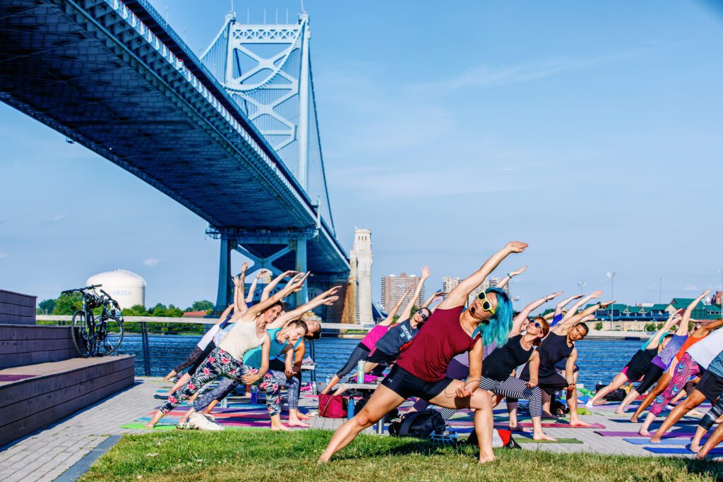 A group of people stretching while doing yoga below the Benjamin Franklin Bridge at Race Street Pier.