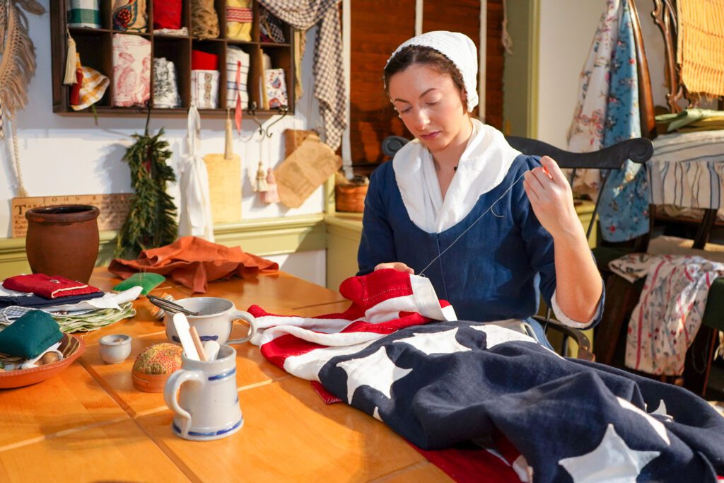 A Betsy Ross impersonator sewing the original American flag at at table.