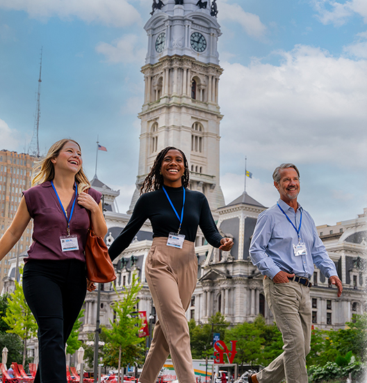 Three people wearing conference badges walk in front of Philadelphia's City Hall.