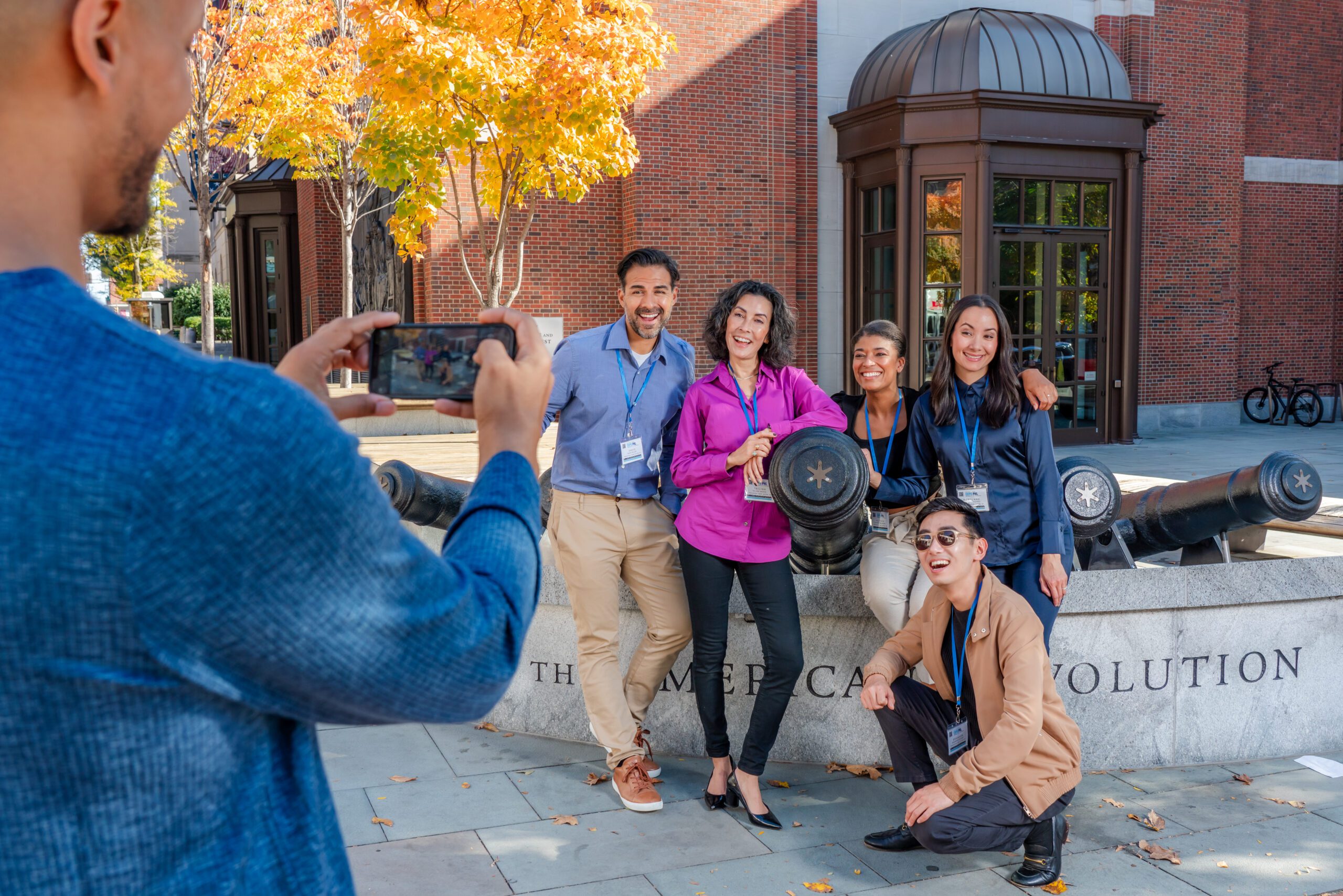 A person taking a photo of a group of five people posing in front of a canon placed outside of the Museum of the American Revolution.