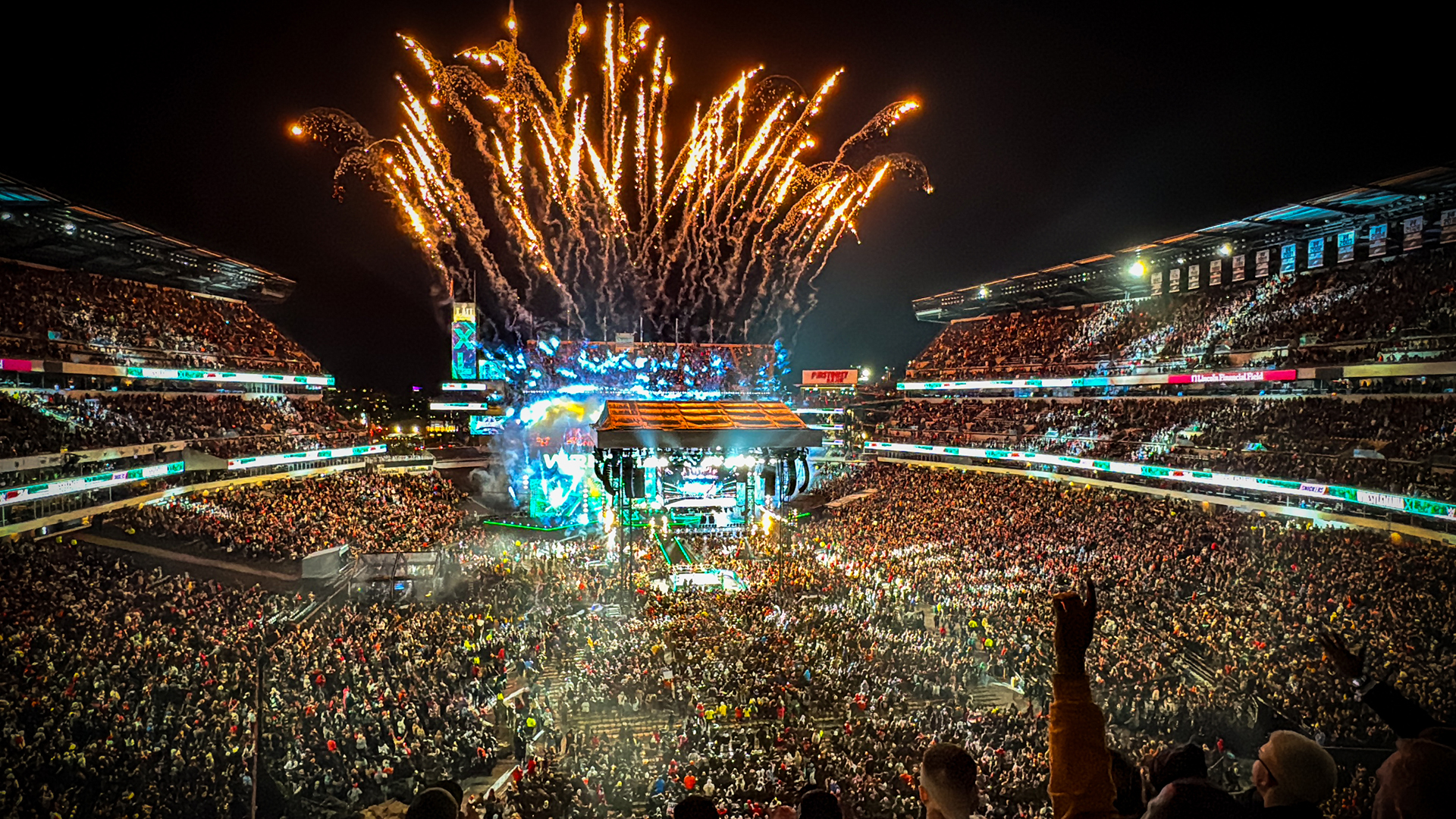 A huge crowd gathers in stands in a stadium with fireworks in the background.