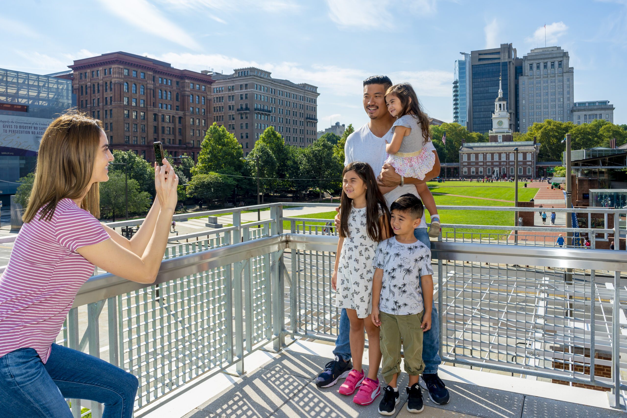 A family taking a photo on the balcony of the Independence Visitor Center in the heart of Philadelphia’s Historic District overlooks Independence Mall, with The President’s House: Freedom and Slavery in the Making of a Nation in the foreground, and, in the distance, Independence Hall.