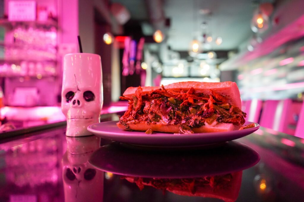 A vegan cheesesteak on the bar of Monster Vegan next to a skull bar glass, both being shined on by a pink neon light