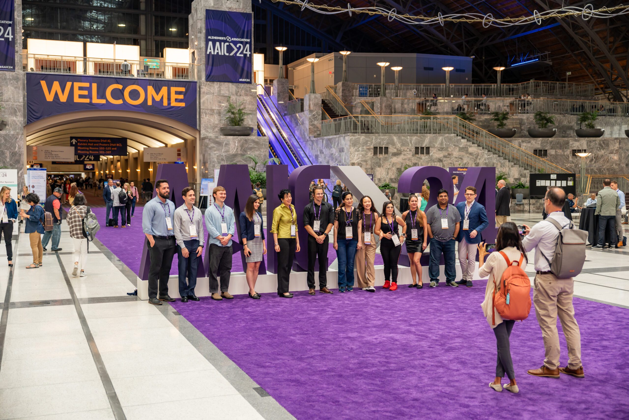 A group of meeting attendees pose in front a AAIC24 in the main hall of the Pennsylvania Convention Center.